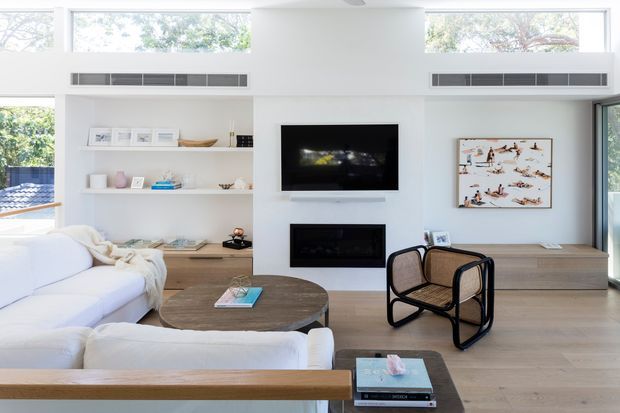 Bright, contemporary living room with white built-in shelving and cabinetry, a central wall-mounted television above a linear fireplace, extensive clerestory and corner glazing, pale timber flooring, and a neutral upholstered sectional sofa. A round timber coffee table and woven black-framed lounge chair add warmth and texture.