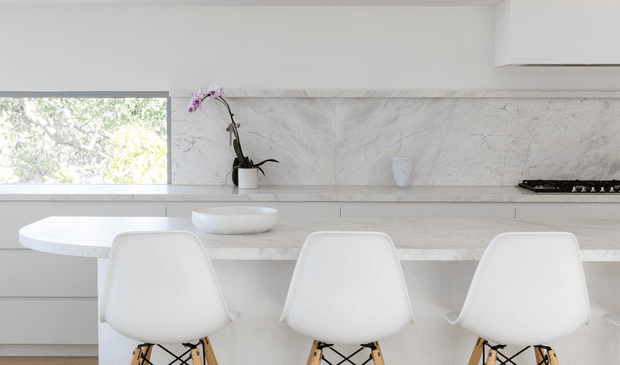 Bright, minimalist contemporary kitchen with a long pale stone island and integrated round white basin, three white molded chairs with timber legs, and a matching marble-look splashback. White cabinetry, a horizontal window, subtle black cooktop, and a potted orchid complete the clean, light-filled space.