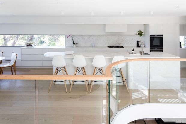 Bright contemporary open-plan kitchen viewed from an adjacent stair or landing, with a large white island, integrated white cabinetry, marble-look backsplash, built-in oven, timber flooring, and white bar stools. A glass balustrade with a curved timber handrail overlooks the stair void in the foreground. Dining furniture and outdoor greenery are visible through the glazing.