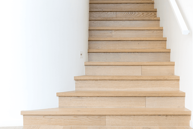 Bright minimalist interior view looking up a straight staircase with pale natural timber treads, risers and side trim. The stairs are set against smooth white walls, with a simple white handrail visible along the right side. The composition is clean and architectural, emphasizing the timber grain and stepped geometry.