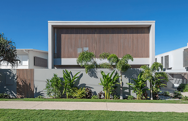 Front exterior view of a contemporary multi-level residence with a crisp white rectangular facade, large recessed upper-level opening, and warm vertical timber screening. Tropical planting and a paved frontage soften the clean geometric architecture under a clear blue sky.