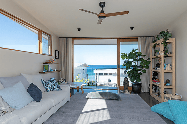 Bright coastal living room with a white sofa, patterned and blue cushions, large windows and sliding glass doors opening to an ocean view. The room features a grey carpet, timber-framed glazing, a ceiling fan, indoor plants, open timber shelving, and a low coffee table. Natural light creates a relaxed, airy atmosphere.