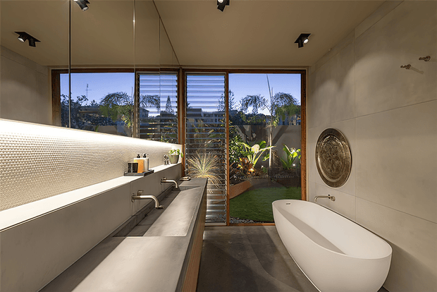 Contemporary bathroom with a freestanding white bathtub, long double vanity, large mirror, textured tiled splashback, and full-height louvred glazing opening toward a planted courtyard. Warm timber-framed windows, concrete-look flooring, and integrated recessed lighting create a refined indoor-outdoor atmosphere.