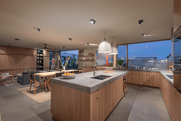 Open-plan contemporary kitchen and living area with extensive warm timber cabinetry, a large central island with a grey stone benchtop, integrated cooktop and sink, pendant lighting, and concrete-look flooring. The adjacent living and dining spaces open toward large glazed doors overlooking a balcony or terrace.