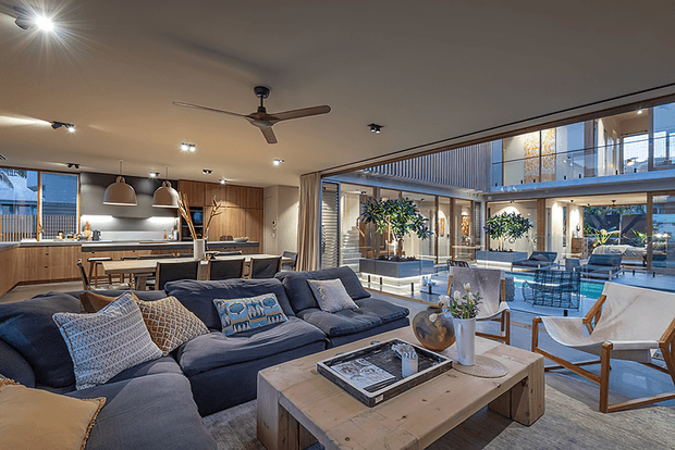 Open-plan contemporary living area with a large navy sectional sofa and timber coffee table, connected directly to a timber-finished kitchen and dining zone. Extensive floor-to-ceiling glazing overlooks a bright internal courtyard or outdoor entertaining area, with a ceiling fan, pendant lights, built-in cabinetry, and layered indoor-outdoor seating.