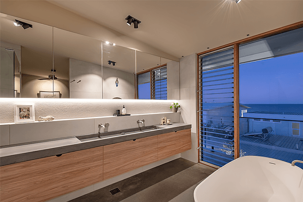 Contemporary bathroom with a long floating timber vanity, dark stone benchtop, twin basins and wall-mounted taps. A large mirrored wall extends above a softly illuminated shelf, while louvred windows and a glazed door open to an ocean-facing view. A freestanding white bathtub occupies the foreground.