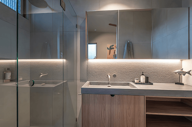 Contemporary bathroom vanity and walk-in shower with a large mirrored wall cabinet, frameless glass shower enclosure, wall-mounted basin, textured mosaic splashback, integrated under-cabinet lighting, and warm timber-look vanity cabinetry. Neutral grey and wood tones create a refined, softly lit interior.