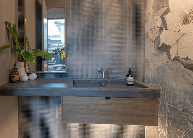 A compact contemporary bathroom vanity with a long grey stone or concrete benchtop, integrated basin and wall-mounted brushed metal tap. The vanity has a timber-look drawer front, while the walls are clad in small square mosaic tiles and a large monochrome botanical mural. A frameless mirror reflects an exterior view and small decorative toiletries sit on the counter.