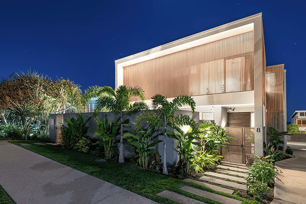 Night-time exterior view of a contemporary multi-level residence with a crisp rectangular facade, large warm-lit upper screening panels, vertical timber-toned slatted detailing, and a recessed ground-floor entry. Tropical landscaping with palms and dense planting borders the concrete driveway and stepping-stone path.