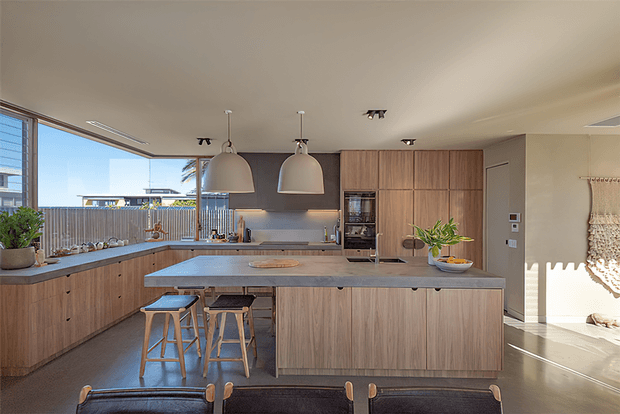 A bright contemporary open-plan kitchen with extensive warm timber cabinetry, a large central island with a pale stone benchtop, integrated appliances, and two pendant lights. Bar stools and dining chairs occupy the foreground, while large windows provide abundant daylight and views of the surrounding urban setting.