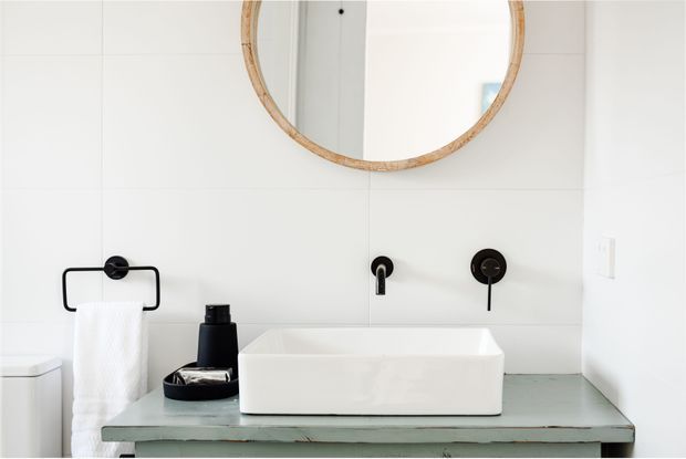 Bright, minimalist bathroom vanity featuring a white vessel basin on a weathered pale green-grey built-in or fixed vanity surface. Matte black wall-mounted tapware and towel rail provide contrast, while a large round timber-framed mirror is mounted above. White tiled walls create a clean, contemporary look.
