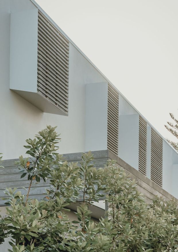 Upward-looking exterior view of a minimalist contemporary building facade, featuring white geometric wall projections, repeated louvred screening, and horizontal grey stone or concrete bands partially screened by mature leafy planting.