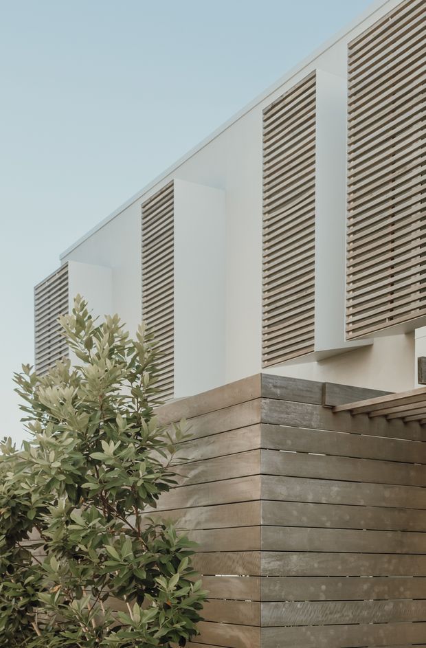 Close-up exterior view of a contemporary multi-level residential facade. White rendered wall volumes are punctuated by tall horizontal timber-look louvred screens, while the lower level features weathered horizontal timber cladding and a projecting timber roof or balcony edge. A leafy shrub partially obscures the foreground, reinforcing the warm modern architectural character.