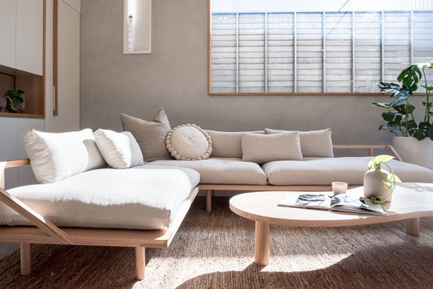 Bright, calm living room with a low-profile L-shaped cream sectional sofa, neutral cushions, round timber coffee table, textured natural-fibre rug, indoor plants, and warm timber-framed built-in cabinetry. Large horizontal windows with blinds provide abundant daylight, while pale plaster walls and timber accents create a refined contemporary interior.