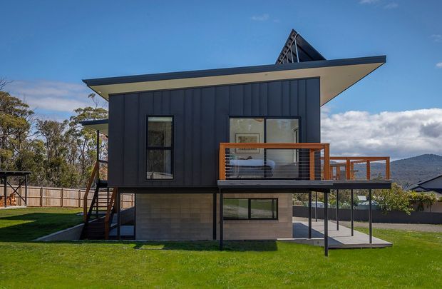 A contemporary elevated two-storey house viewed from the rear garden. The upper level has dark vertical cladding, expansive eaves, a projecting balcony with timber-toned railings, and a steep triangular roof element visible above. The lower level combines pale masonry or concrete block walls with extensive glazing and an open covered area. The house sits on a landscaped suburban site with lawn, fencing, neighbouring homes, and wooded hills in the background.