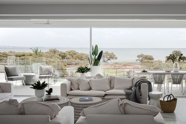 Bright open-plan living room with a soft neutral palette, large white sofas, woven round coffee tables, indoor plants, and expansive floor-to-ceiling glazing overlooking a coastal landscape and water. A dining area is visible behind the seating, creating an airy, relaxed contemporary interior.