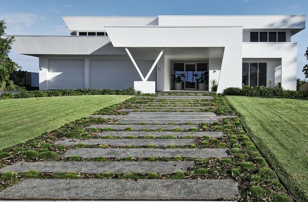 Street-facing view of a large, angular white modern house with a flat roof, projecting geometric roof planes, deep entry canopy, extensive glazing, and an integrated double garage. A broad landscaped front lawn and stepping-stone driveway lead to the recessed entrance.