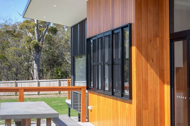 A contemporary timber-clad residential exterior viewed from a raised deck or patio. The facade combines warm vertical timber boards with dark charcoal cladding and dark-framed windows and doors beneath a broad flat roof overhang. A simple outdoor table and bench occupy the foreground, with a fenced, wooded setting beyond.