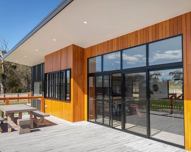 Contemporary single-storey building exterior with warm vertical timber cladding, extensive black-framed glazing, and a broad flat roof overhang. The image shows a timber deck with outdoor picnic-style seating, while the glazed interior reveals a dining or living area. The setting appears leafy and residential, with a bright blue-sky backdrop.