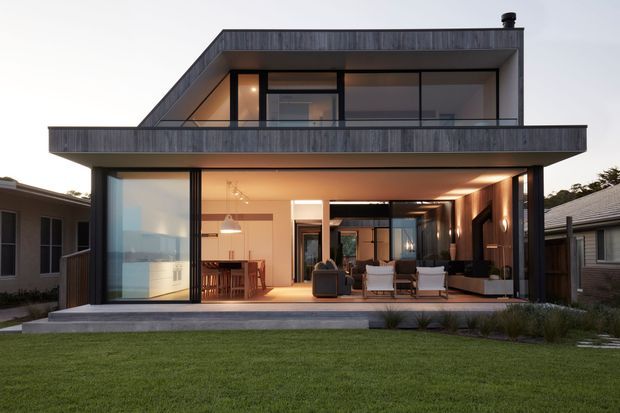 Contemporary two-storey residential home viewed from the rear garden, featuring a strong cantilevered upper level, dark timber or composite cladding, broad balconies, and extensive floor-to-ceiling glazing. The open ground floor connects a warm, softly lit living and dining area to the lawn, while the upper level has a private balcony and glazed bedroom or living space. The design has a flat roof, clean rectilinear lines, and a restrained dark-and-warm palette.