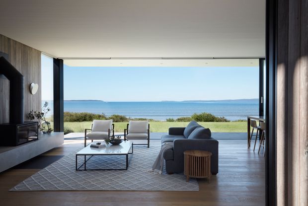 A refined coastal living room with an open-plan, full-width glazed opening overlooking a lawn, dunes and the ocean. The room features a deep grey sectional sofa, two light upholstered lounge chairs, a low black-framed coffee table, a textured area rug, timber flooring, vertical timber wall panelling, and a minimalist built-in media wall. A dining area is partially visible to the right. The interior is calm, airy and contemporary, with strong indoor-outdoor connection and expansive ocean views.
