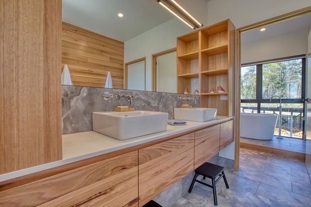 Warm contemporary bathroom with a long timber vanity featuring two vessel basins, open timber shelving, large mirrors, and a grey stone-pattern splashback. Timber wall panelling and framed openings lead toward a freestanding bathtub and glazed outdoor view.