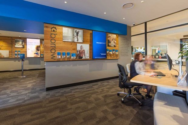 A contemporary commercial reception or customer service area with a prominent blue feature wall, timber display panels, branded signage, integrated brochure shelving, and a long light-coloured service counter. The space has extensive black-framed glazing, carpet tile flooring, office chairs, workstations, and bright recessed lighting.