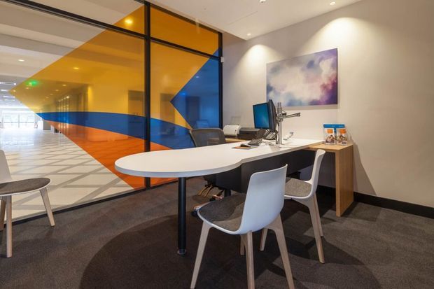 A modern commercial office meeting or collaboration area with a white oval table, computer workstation, visitor chairs, and a wood-finished desk. The space has grey carpet, light neutral walls, recessed ceiling lighting, and a striking floor-to-ceiling black-framed glass partition with bold translucent orange and blue geometric graphics. An abstract artwork hangs on the wall, while a bright open circulation area is visible beyond the glazing.