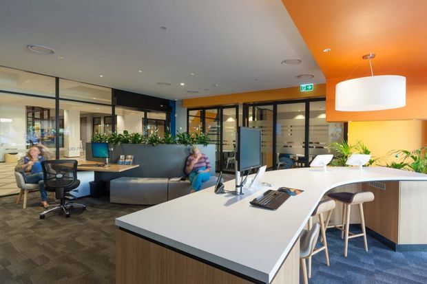 Bright contemporary commercial office interior with an open-plan reception or collaborative workspace. A large white island workbench with integrated power and computer equipment dominates the foreground, accompanied by bar stools. Behind it are desks, office chairs, indoor plants, glass-partitioned meeting rooms, and a curved grey upholstered bench. The space uses orange and white ceiling planes, dark-framed glazing, wood-look cabinetry, patterned carpet, and large pendant lighting.