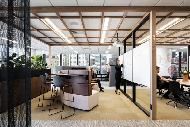 A contemporary open-plan commercial office interior with glazed meeting rooms, desks, office chairs, a communal upholstered seating and work area, and a large writable whiteboard. The space features a warm timber ceiling grid and columns, carpet flooring, black-framed glass partitions, integrated linear lighting, and views toward a dense urban skyline. The atmosphere is bright, professional, and collaborative.