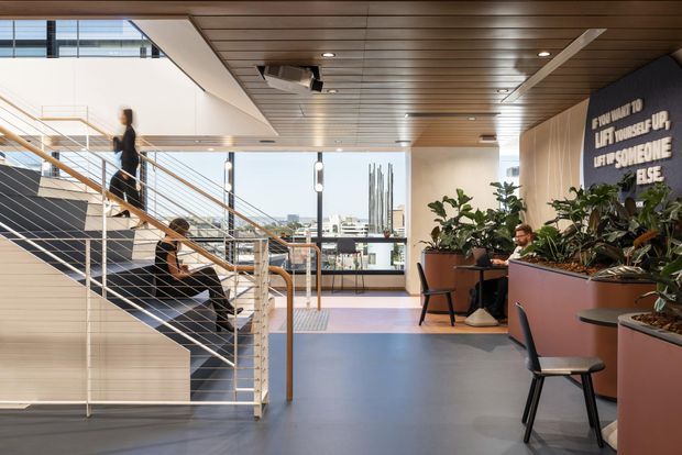 A bright contemporary commercial interior with an open stairway, slim metal balustrades, extensive glazing, and a landscaped planter-lined lounge or circulation area. The space has a blue-grey floor, warm timber slatted ceiling, pale walls, integrated lighting, and casual black seating. A visible urban skyline and motivational wall lettering reinforce the workplace or public-building setting.