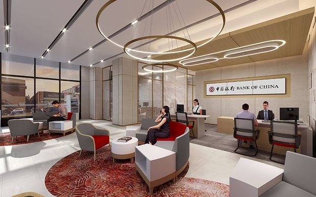 A contemporary bank branch interior with a reception and customer service area. The space features a bright open-plan layout, white and light grey finishes, marble-look wall surfaces, timber ceiling accents, glass partitions, sculptural circular LED pendant lights, red and grey lounge seating, and patterned red area rugs. The Bank of China sign is prominently displayed behind the service desk.