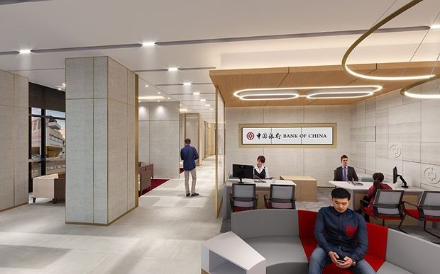 A contemporary bank branch interior with a spacious customer service and waiting area. The image shows reception desks, computers, staff and visitors, a Bank of China sign, pale stone-look flooring, large textured feature columns, glass partitions, and sculptural linear pendant lighting. The fit-out uses a restrained palette of white, light grey, beige, timber and red accents.
