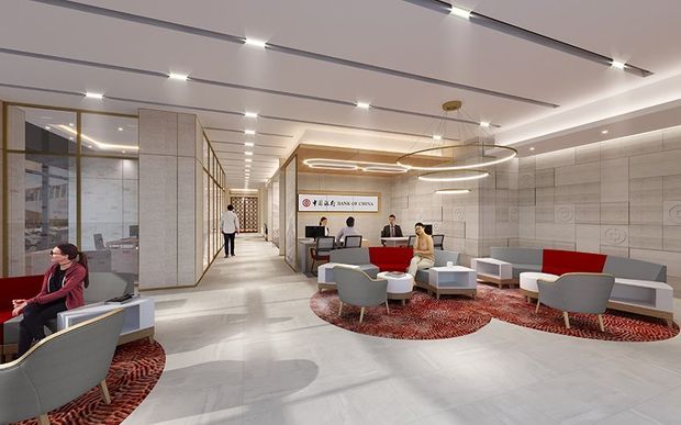 A bright, contemporary bank lobby and customer service area with pale stone-look wall and floor finishes, glazed partitions with metal framing, linear recessed ceiling lighting, and sculptural circular pendant lights. A reception and teller counter sits at the rear, while lounge seating is arranged on large red patterned rugs. The space has a spacious, polished commercial interior with clear circulation and a refined corporate atmosphere.
