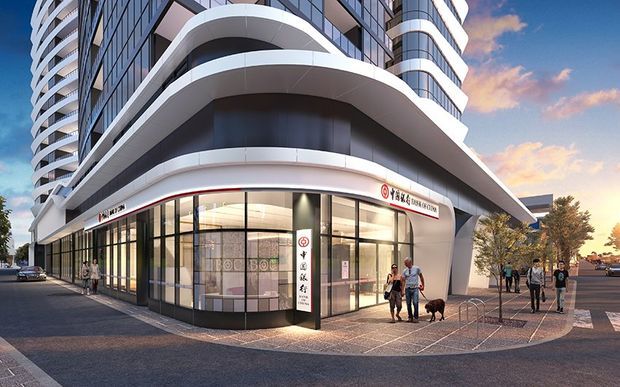 A street-level view of a modern multi-storey mixed-use building with a curved white architectural canopy, rounded balconies, extensive dark-framed glazing, and a ground-floor commercial tenancy with Chinese signage. The building occupies a landscaped urban pedestrian frontage with paving, street trees, and surrounding high-density development.