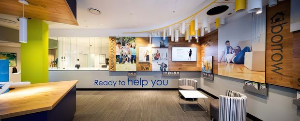 A contemporary commercial reception and waiting area with a prominent service counter, branded wall graphics, illuminated pendant lighting, timber feature ceiling and wall panels, and striped lounge seating. The space has a polished, welcoming workplace or showroom character with a large reception desk and visible signage.