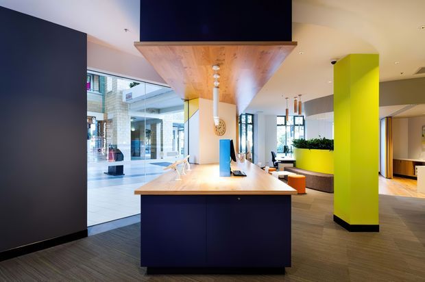 A contemporary commercial interior lobby or reception area featuring a central island counter with dark built-in cabinetry and a light timber benchtop. A timber-clad suspended canopy supports a large black display or screen above the counter, with pendant lights underneath. The space has dark carpet flooring, extensive storefront glazing, bright lime-green architectural columns and planter joinery, and casual seating in the background.