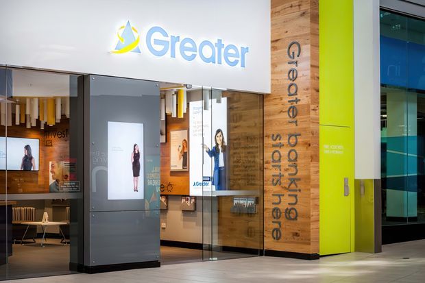 Contemporary commercial bank branch interior and storefront, featuring a prominent Greater Bank logo, bright lime-green branded wall, timber feature cladding with vertical lettering, glass entry doors, illuminated display panels, and a grey tiled floor. The space has a polished retail fit-out with strong branded graphics and extensive glazing.