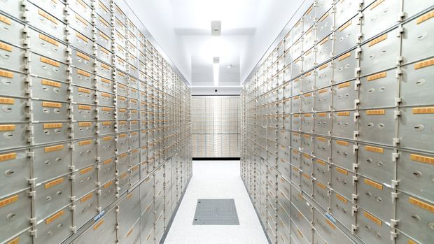 A bright, narrow institutional storage corridor lined on both sides with extensive floor-to-ceiling banks of small metal lockers or secure storage compartments. The space has white walls and ceiling, evenly spaced recessed lighting, a pale hard-wearing floor with a central access panel, and a clean, highly repetitive utilitarian appearance.