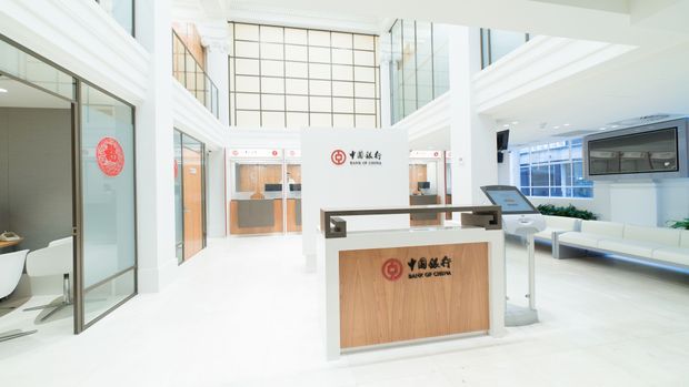 Bright, double-height commercial bank interior with a central timber-front reception or information desk, white architectural finishes, extensive glass partitions, a large gridded feature window, and customer seating along the right side. The space has a clean, contemporary institutional design with abundant daylight and polished, uncluttered surfaces.