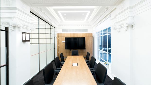 Bright, elegant conference room with a long light-wood meeting table surrounded by black upholstered chairs. The room features ornate white classical wall mouldings and a decorative coffered ceiling, contrasted with black-framed glass partitions on one side and large blue-tinted windows on the other. A timber feature wall with a mounted television forms the focal point.