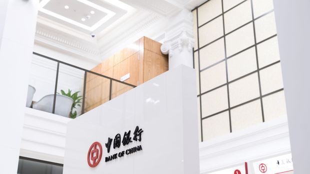 Bright interior view of a Bank of China branch lobby, featuring a prominent white reception or signage wall with the bank logo, black-framed gridded glazing, a partial mezzanine with glass balustrade, warm timber wall panelling, and ornate white classical mouldings and ceiling detailing.