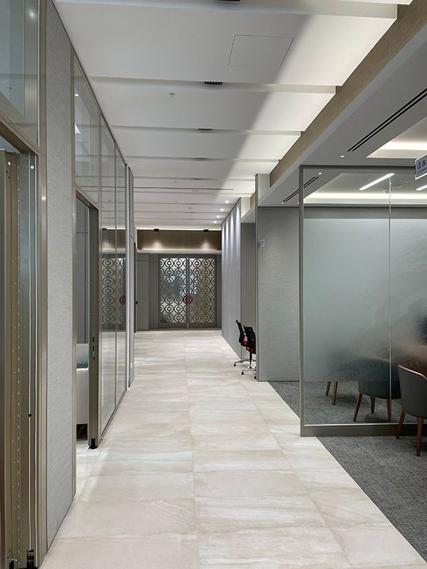 A bright, modern commercial office corridor with pale large-format tiled flooring, glass-partitioned meeting rooms and offices, grey textured walls, and a white recessed ceiling with linear lighting. Decorative patterned metal screens feature at the far end, while a small seating area is visible through the glazing on the right.