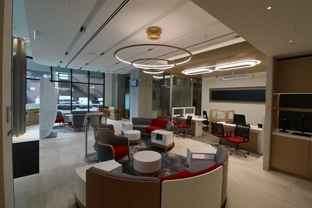 A contemporary commercial interior resembling a reception and informal waiting lounge, with curved upholstered seating, small round and rectangular coffee tables, workstations, and a built-in media or display joinery wall. Large glazed windows line the left side, while layered circular pendant lights, pale flooring, dark glass partitions, and warm timber cabinetry create a refined office or hospitality-like setting.