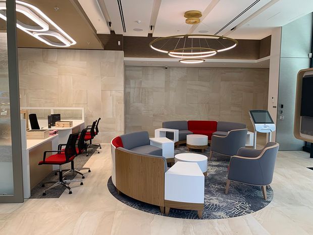 A refined contemporary commercial reception or waiting area with a circular arrangement of upholstered lounge chairs and small white tables on a patterned round rug. An adjacent workstation zone with red-and-black office chairs is visible on the left. The space features pale stone-look wall and floor finishes, dark timber-toned ceiling and wall accents, integrated linear lighting, and large sculptural circular pendant fixtures.