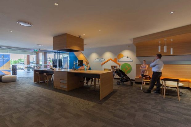 A contemporary commercial hospitality or workplace lounge with an open-plan timber island counter, integrated seating, pendant lights, and a long built-in banquette along the rear wall. The space has dark patterned carpet, warm timber cabinetry and wall panelling, recessed ceiling lights, and large glazed entry doors. Colourful graphic artwork and several people with a stroller are visible, suggesting a welcoming public or shared amenity area.