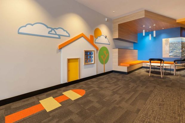 A brightly coloured contemporary commercial interior, likely a children's or family-oriented waiting and activity area. The space features playful house, cloud, tree and sun wall graphics, a small yellow doorway, patterned carpet with orange and yellow geometric floor accents, vivid blue and orange feature walls, and integrated timber-look ceiling and wall joinery. A small meeting or seating area is visible at the rear.