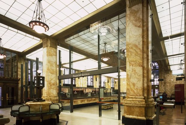 Interior view of an ornate financial-services hall, featuring monumental veined stone-clad columns with classical bases and capitals, dark timber detailing, extensive mirrored glazing, a luminous gridded coffer-like ceiling, and suspended decorative chandeliers. Financial service counters and desks occupy the central space, with a bank-style clock or information display in the foreground. The overall character is grand, historic, and richly detailed.
