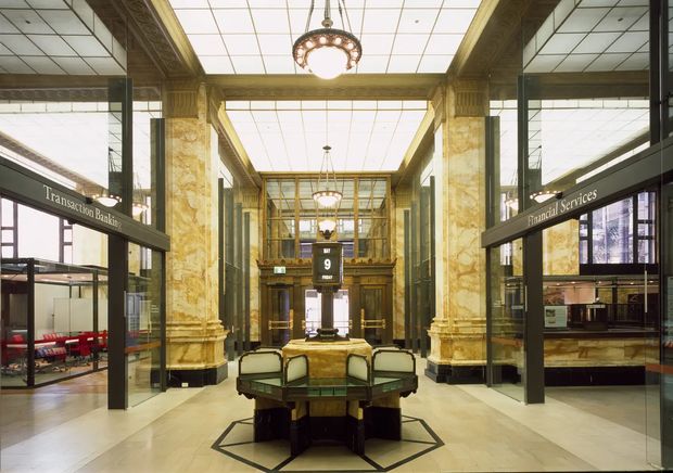 Grand historic bank interior with a symmetrical central lobby, ornate veined stone-clad columns, dark timber and bronze-toned detailing, extensive glazed teller and service counters, a coffered luminous ceiling, and decorative pendant chandeliers. A central polygonal customer service or transaction desk anchors the spacious hall.