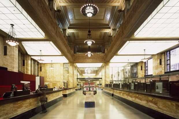 Grand historic commercial interior resembling a restored railway station or civic hall, with a long symmetrical concourse, ornate carved and coffered ceiling, decorative chandeliers, polished stone or marble counters and flooring, monumental columns, and extensive glazed service or retail fronts. The space combines heritage detailing with modern illuminated ceiling panels and contemporary commercial fittings.